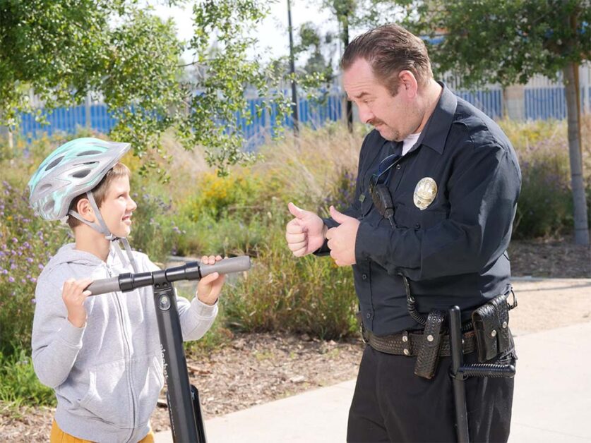 A policeman giving thumbs to a boy with electric scooter and a helmet.